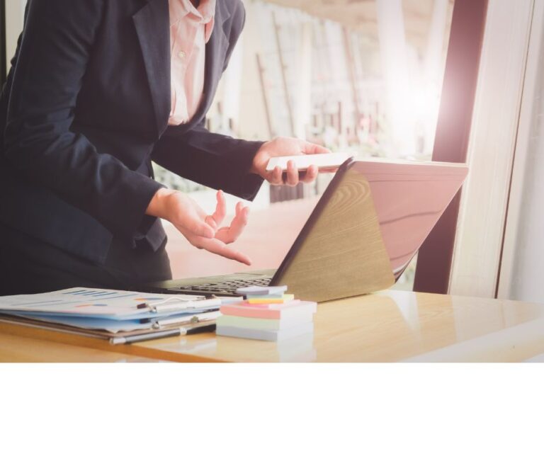 A businessperson in a suit standing by a desk with a laptop, gesturing while holding a phone, with documents and sticky notes on the desk in a well-lit office setting.