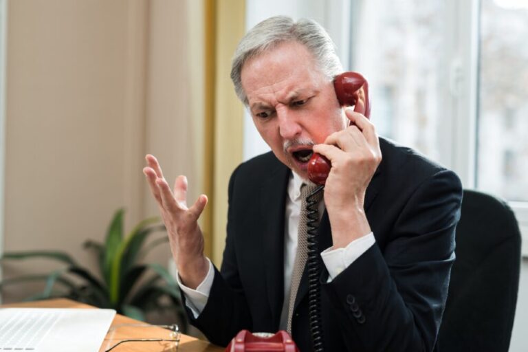 A frustrated man in a suit holding a red corded phone, gesturing with his hand while speaking, seated at a desk with a laptop in a brightly lit office.